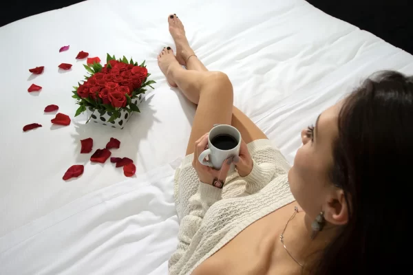 Donna su un letto bianco con una tazza di caffè e un bouquet di rose rosse.