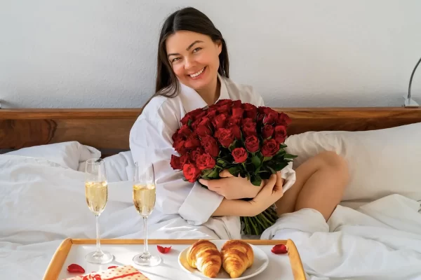Donna sorridente seduta sul letto con un mazzo di rose rosse e una colazione speciale su un vassoio.