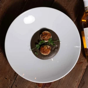 Capesante su crema scura, guarnite con erbe fresche, in un piatto bianco su tavolo di legno.