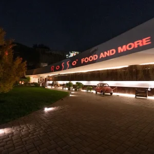 Edificio moderno illuminato di notte con una scritta "Rosso: Food and More" e un'auto classica rossa parcheggiata davanti.