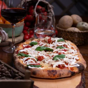 Pizza gourmet con basilico, pomodorini e formaggio accanto a bicchieri di vino rosso su un tavolo rustico.