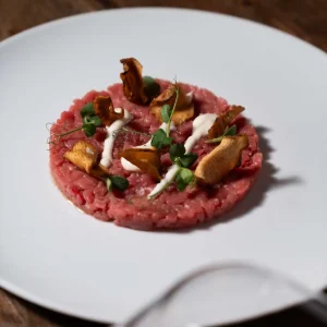 Tartare di carne con funghi, erbe e salse decorative su un piatto bianco.